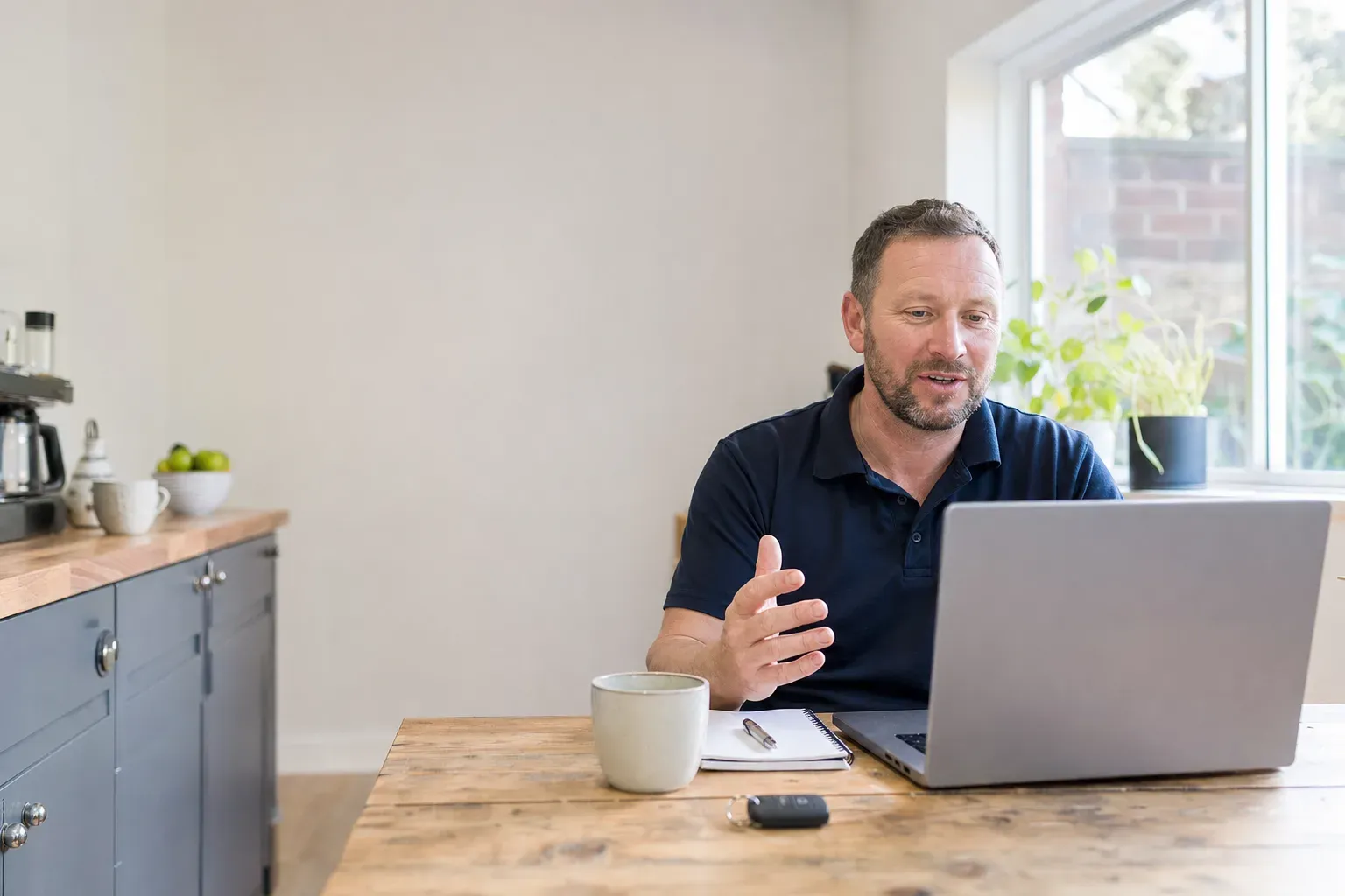A trades business owner on a video call at his kitchen table, notepad and coffee beside the laptop